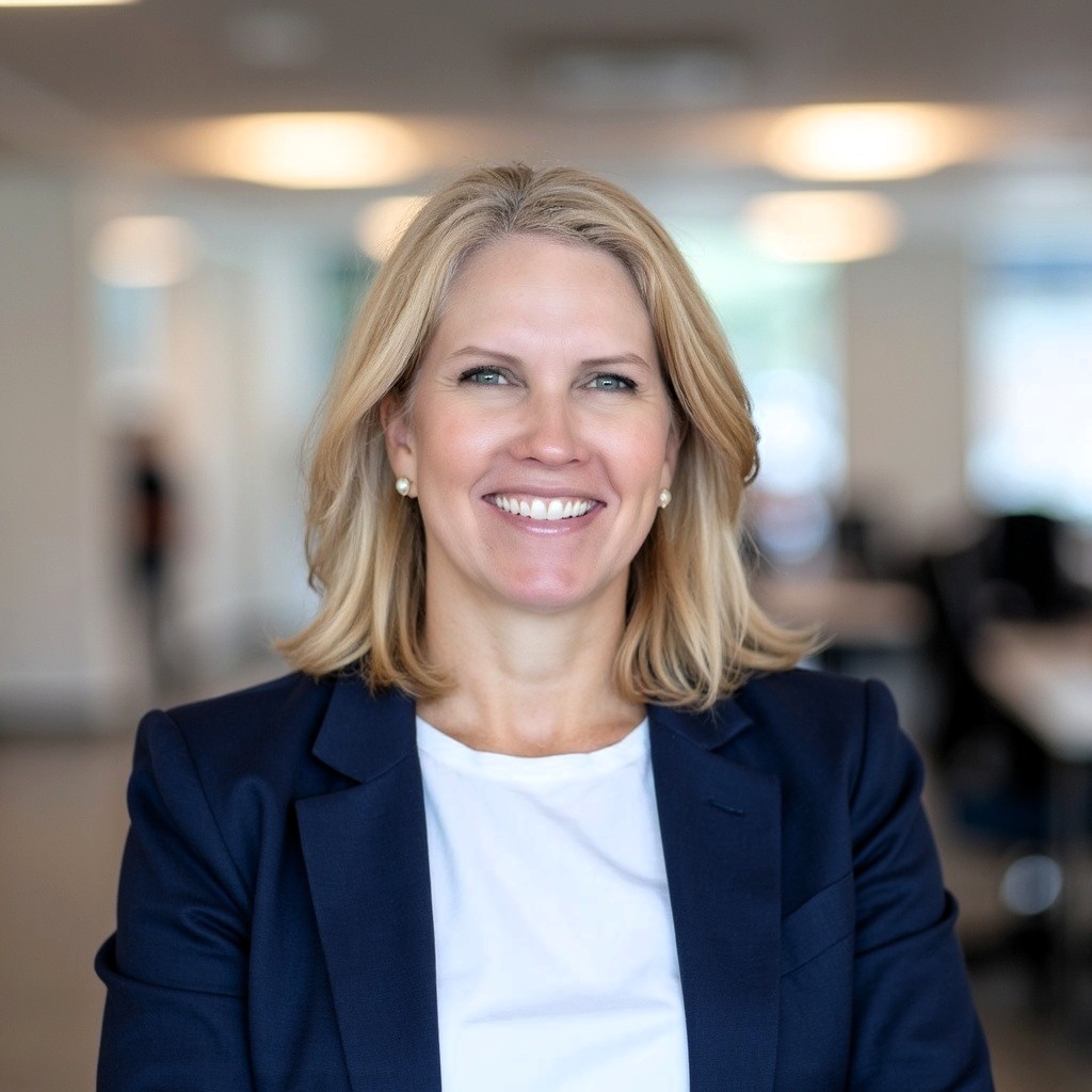 A smiling woman with blonde hair, identified as Lori Fitzsimon, wears a navy blazer and white shirt as she stands in a modern, brightly lit office setting.