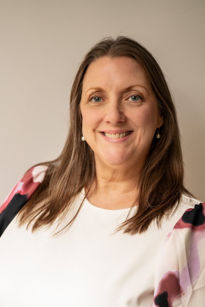 Susan Nonhof, a woman with long brown hair and a warm smile, is wearing a white top with pink and black patterned sleeves, standing against a plain light-colored background.