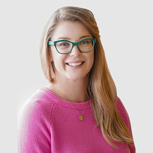 A smiling woman with long blonde hair wearing green glasses, a pink sweater, and a gold necklace, posing in front of a plain light-gray background, showcases the sponsor benefit of positive brand representation.