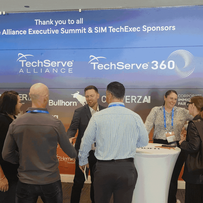 A group of five professionals greet each other and shake hands in front of a TechServe Alliance and TechServe 360 sponsor banner, highlighting the Sponsor Benefit at a business event.