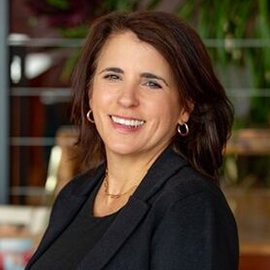Training Series 2 A woman with shoulder-length brown hair, wearing a black blazer and gold hoop earrings, smiles at the camera in an indoor setting with plants in the background, ready to lead a Training Series.