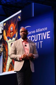 Leading Through Upheaval: Insights from the 2025 TechServe Executive Summit 2 A man in a suit speaks on stage at the Executive Summit, with a comic-style illustration of a football player displayed on a screen behind him.
