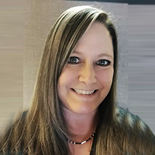 Erica Scoville, a woman with straight, long brown hair and blue eyes, smiles at the camera. She wears a beaded necklace and a black top, set against a neutral, softly blurred background.