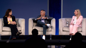 Three people sit in white armchairs on a stage, engaged in a panel discussion. The background is blue, and a silhouette of an audience member is visible in the foreground.