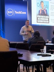 Leading Through Upheaval: Insights from the 2025 TechServe Executive Summit 5 A man stands and speaks in front of an audience at a SIM TechExec event. Behind him, a screen displays a slide about OpenAI launching an AI-powered hiring platform by mid-2025.
