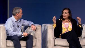 A man and a woman sit on stage in armchairs, engaged in conversation. The man holds papers and listens attentively, while the woman gestures with her hands as she speaks. Both are dressed in business casual attire, with a blue background behind them.