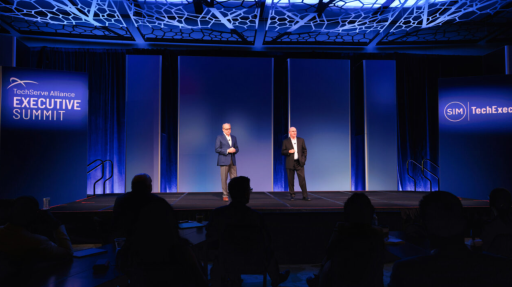Leading Through Upheaval: Insights from the 2025 TechServe Executive Summit 1 Two men in suits stand on a stage under blue lighting at the TechServe Alliance Executive Summit. Audience members are silhouetted in the foreground, and event banners are displayed on either side of the stage.