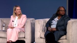 Two women sit on a light-colored couch against a blue background. One wears a light pink suit and looks left, while the other wears a dark suit and looks right. Both appear attentive and engaged in a panel discussion.