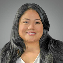 A woman with long, dark hair smiles at the camera. She is wearing a light-colored blouse and a dark blazer, with a neutral gray background behind her.