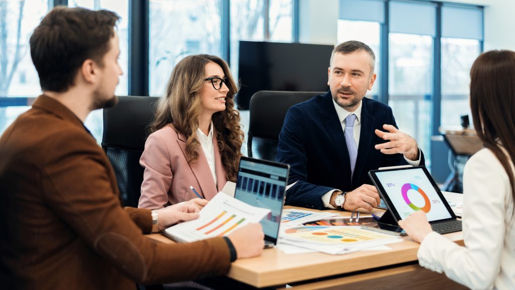 Four professionals sit at a conference table with laptops and charts, discussing business insurance graphs and data. Two men and two women are engaged in conversation in a modern office setting with large windows.