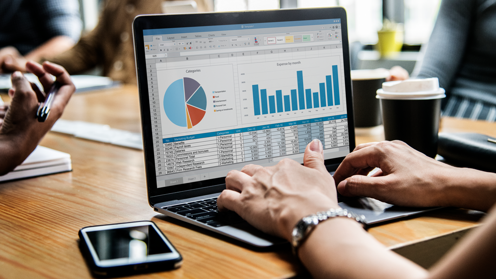 A person works on a laptop displaying spreadsheets with a pie chart and bar graph, possibly analyzing Business Insurance data. Others sit nearby at a table with papers, a coffee cup, and a smartphone.