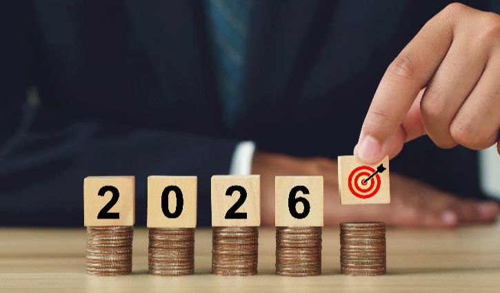 A person in a suit arranges wooden blocks on stacks of coins spelling 2026, with the last block showing a target symbol, symbolizing firm optimization and financial goals in a competitive marketplace for the year 2026.