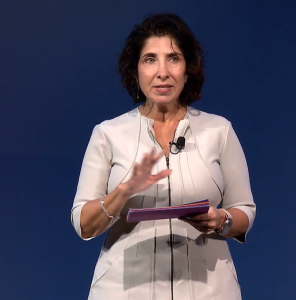 What Today’s IT Staffing Buyers Really Want: How Top Decision-Makers Evaluate, Select, and Trust Their Partners 2 A woman with curly dark hair, wearing a light gray dress, stands against a blue background, speaking and gesturing with her left hand while holding a folder and wearing a microphone clipped to her dress.