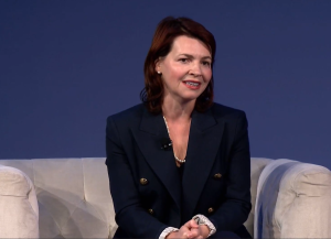 What Today’s IT Staffing Buyers Really Want: How Top Decision-Makers Evaluate, Select, and Trust Their Partners 4 A woman with shoulder-length brown hair, wearing a dark blazer and a pearl necklace, sits on a light-colored sofa against a plain blue background, smiling and speaking.