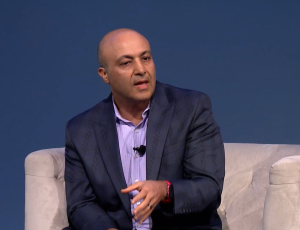 What Today’s IT Staffing Buyers Really Want: How Top Decision-Makers Evaluate, Select, and Trust Their Partners 3 A bald man in a dark checked suit and light purple shirt speaks while sitting on a light-colored couch against a plain blue background, gesturing with his right hand.