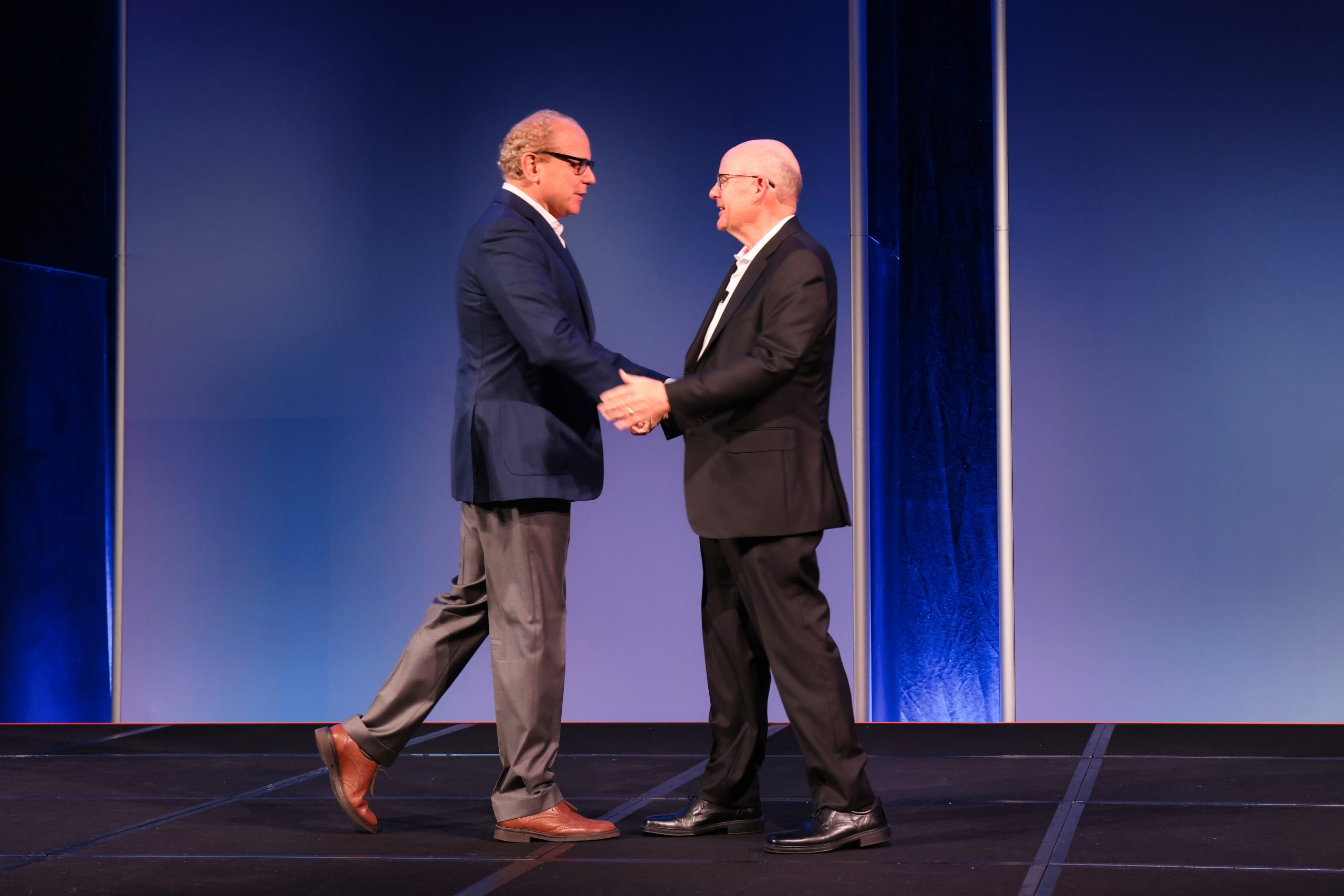 Executive Summit 6 Two men in business attire shake hands and smile at each other on a stage with a blue background, suggesting a formal or professional event.