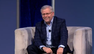 What Today’s IT Staffing Buyers Really Want: How Top Decision-Makers Evaluate, Select, and Trust Their Partners 5 A man wearing glasses, a blue shirt, and a plaid blazer sits smiling on a light-colored sofa against a blue background, with a microphone clipped to his shirt.