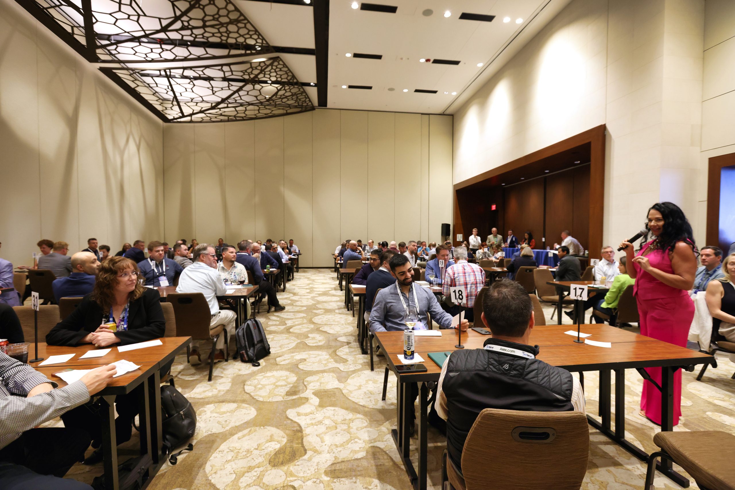 Executive Summit 11 A large conference room with many people seated at tables in pairs, engaging in discussions. A woman in a bright pink outfit stands at the front with a microphone, addressing the attendees. The room has high ceilings and modern decor.