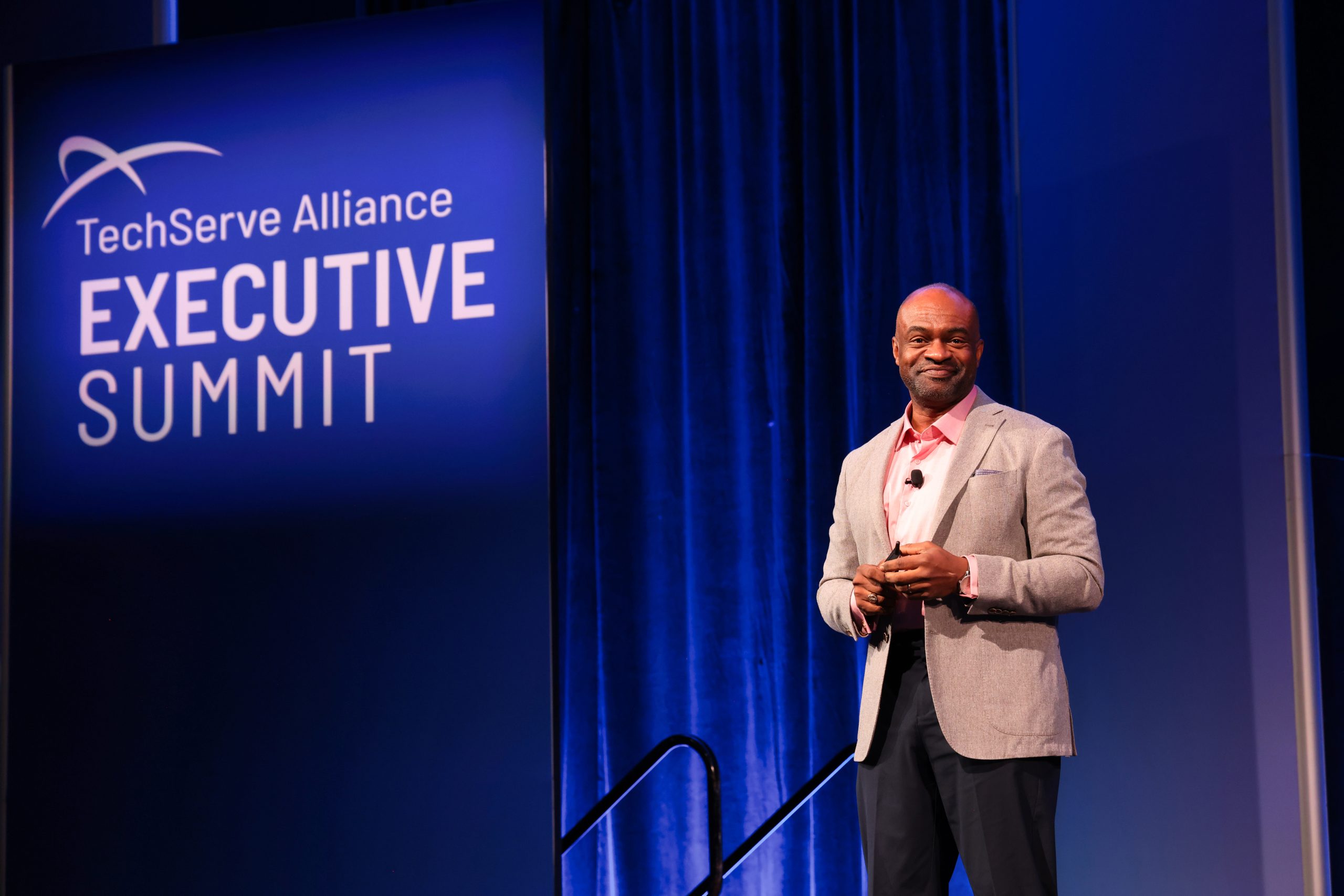 Executive Summit 9 A man in a gray blazer and pink shirt stands on stage, smiling, at the TechServe Alliance Executive Summit. A large event sign is displayed on a blue background behind him.