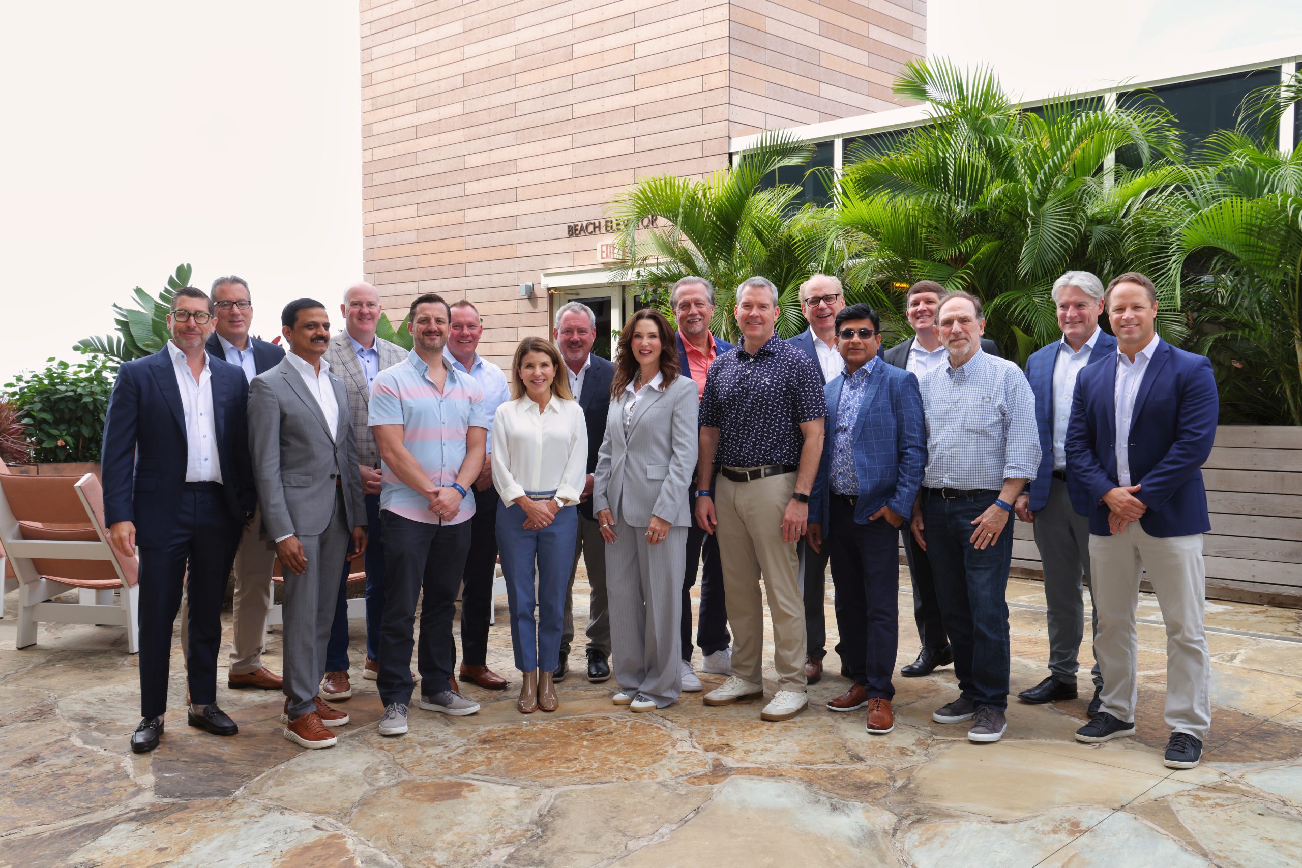 Executive Summit 10 A group of 17 professionally dressed adults, both men and women, stand together outdoors on a sunny day in front of a building and lush green plants, smiling at the camera.