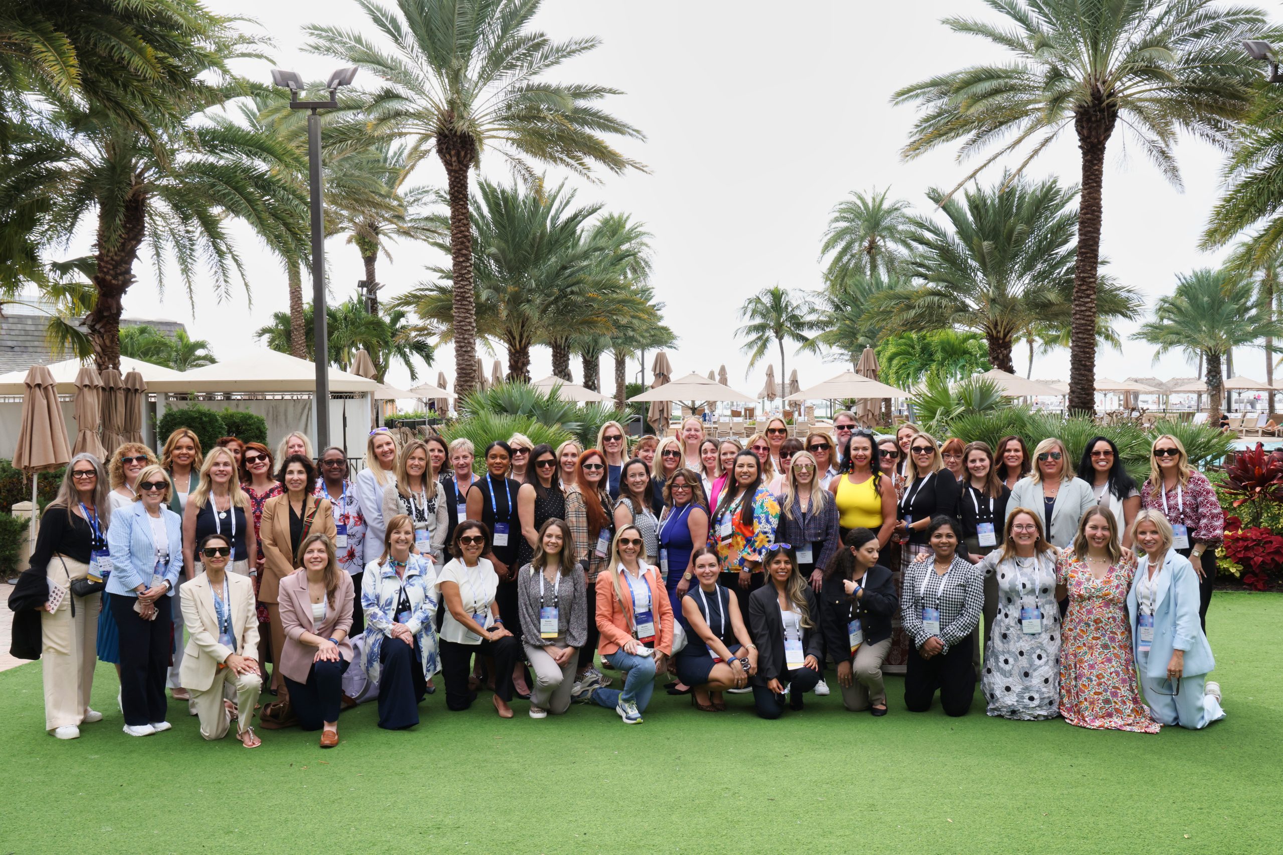 Executive Summit 12 A large group of women pose together outdoors on green grass, surrounded by palm trees and tropical plants, with a resort-like setting in the background. Most are smiling and dressed in business or casual attire.