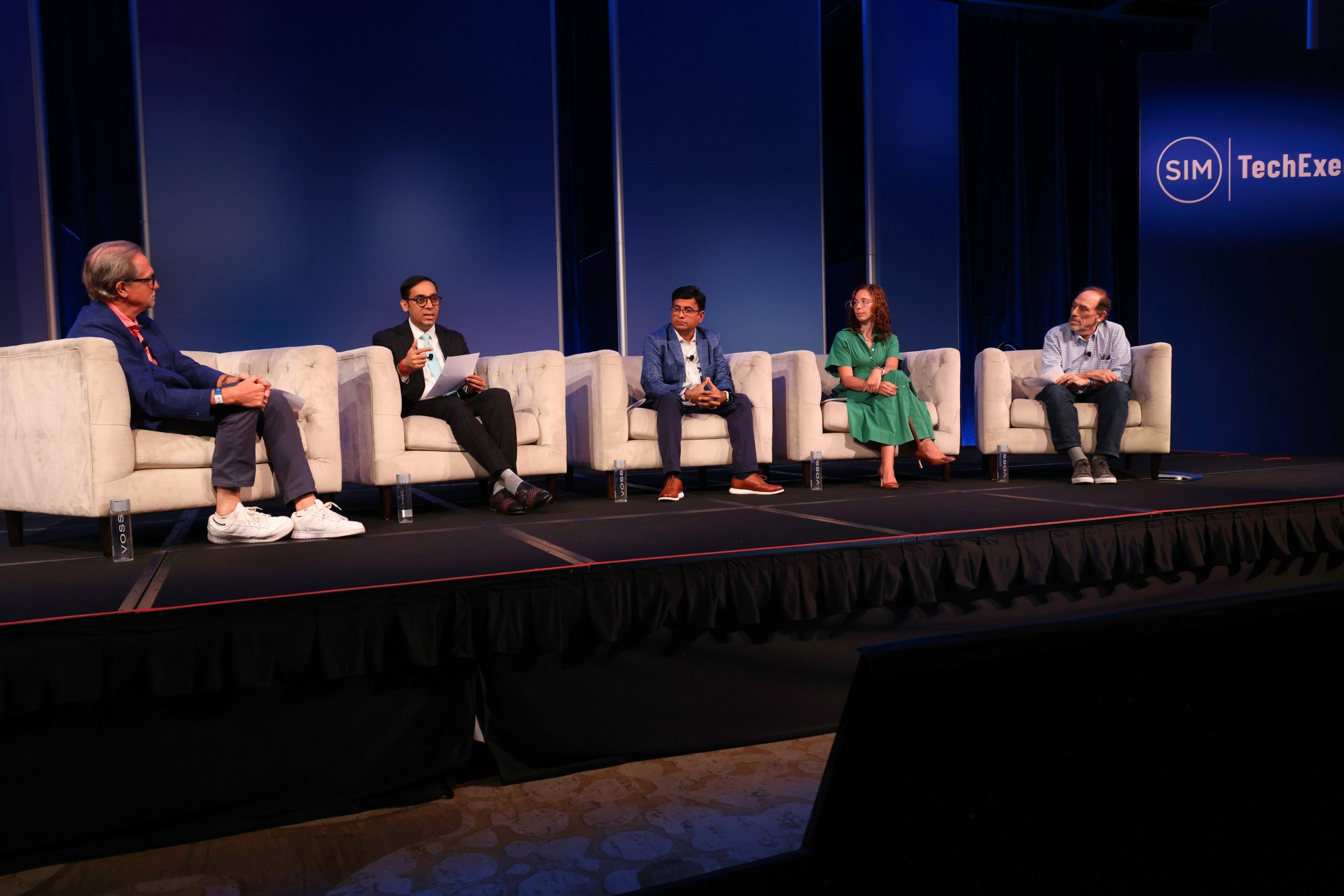 Executive Summit 13 Five people sit on stage in armchairs during a panel discussion at a tech event. Four men and one woman face the audience, with a blue backdrop displaying “SIM” and “TechEx.”.