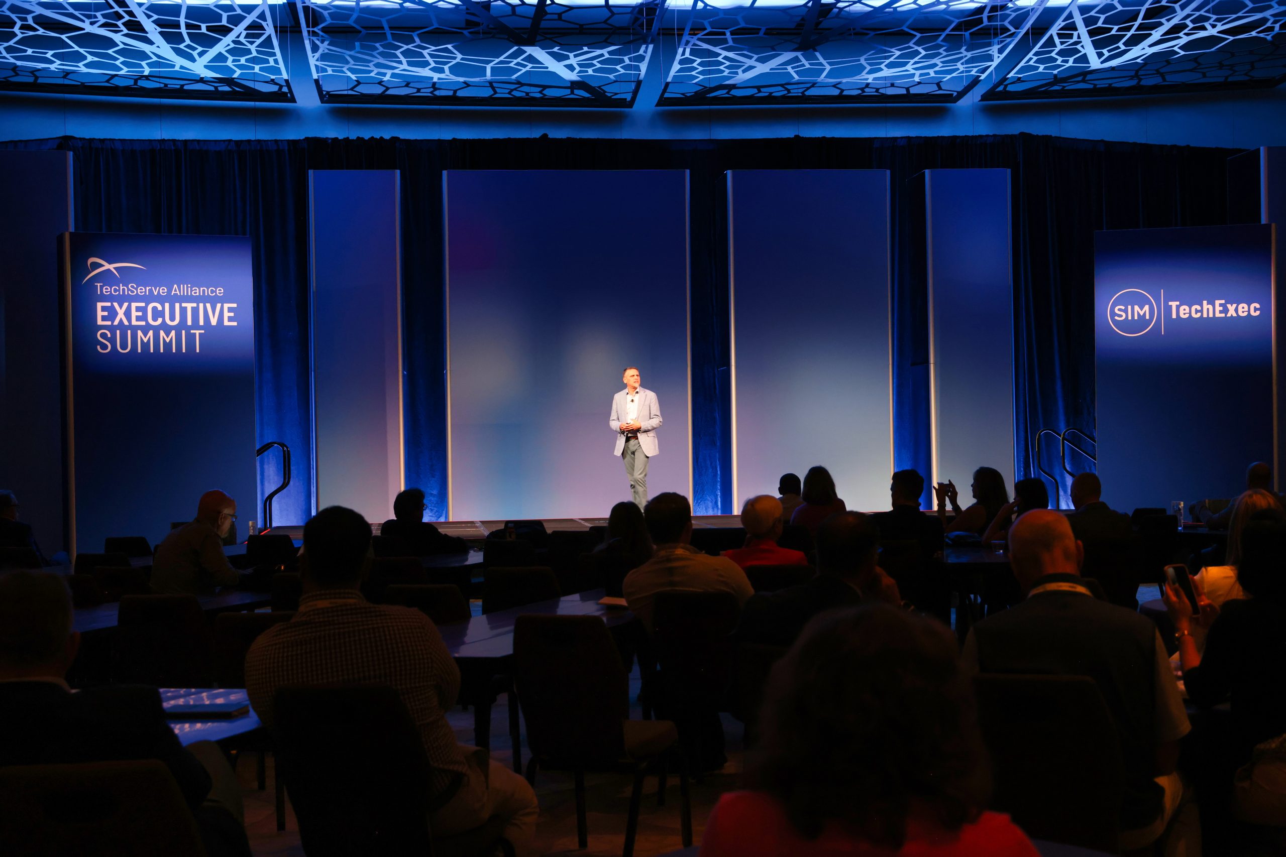 Executive Summit 14 A speaker stands on a stage at the TechServe Alliance Executive Summit, addressing an audience seated at round tables in a dimly lit conference room with blue lighting and backdrop.