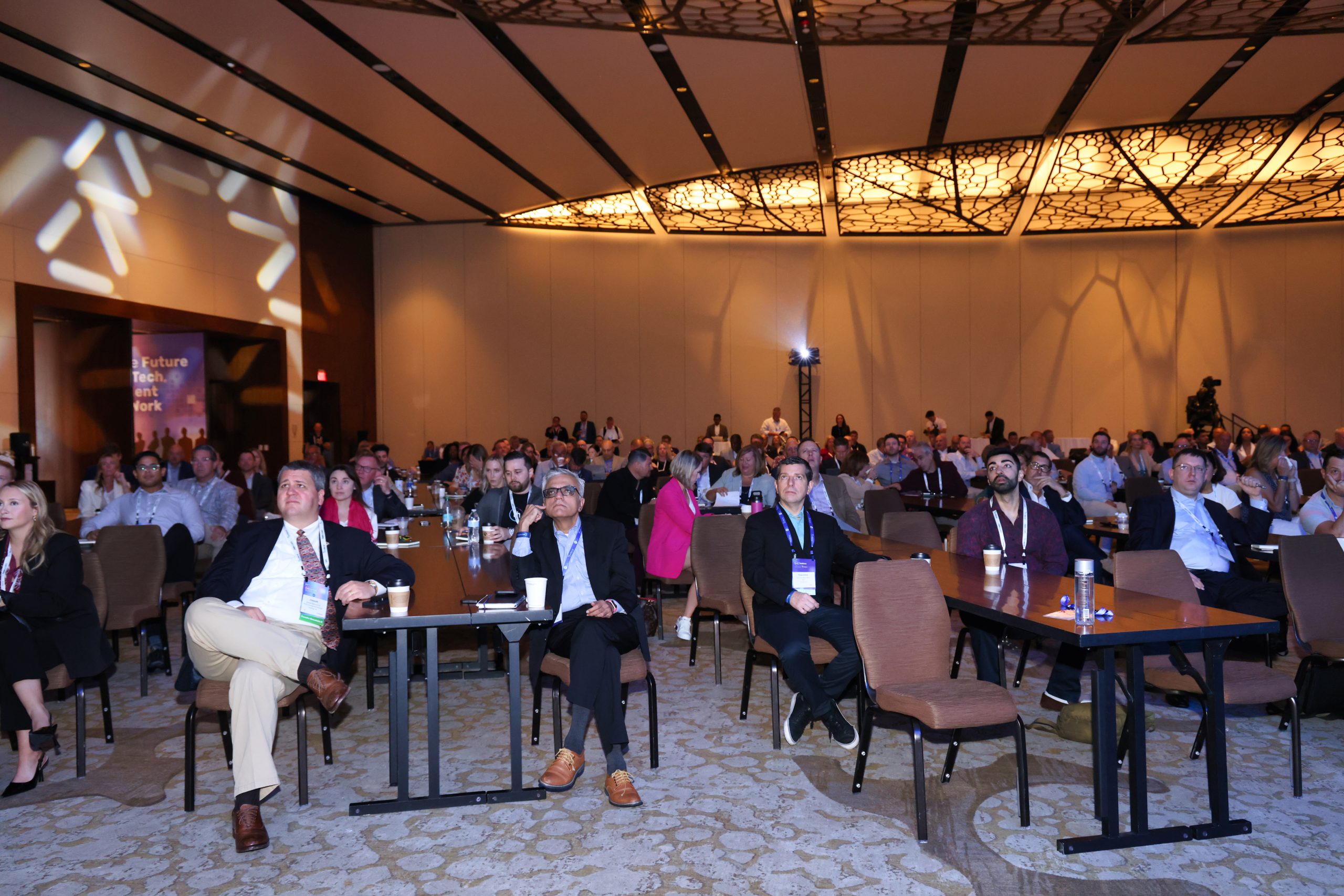 Executive Summit 7 A large group of people sits attentively in a conference room with modern lighting and ceiling design, facing the front where a presentation or event is taking place. Some tables have water bottles and papers.
