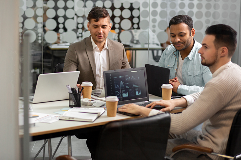 Three men sit at a table in a modern office, working on laptops with charts visible—an active scene of collaboration as they discuss RPO Readiness. Papers, coffee cups, and office supplies hint at a focused talent solutions meeting.