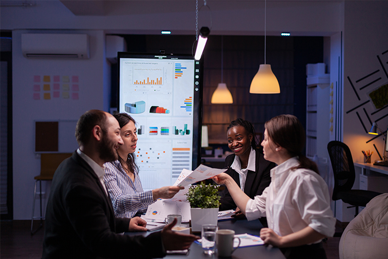 Four colleagues in business attire sit around a table in an office, discussing market documents and charts. A screen behind them displays graphs and data. They appear engaged and collaborative.