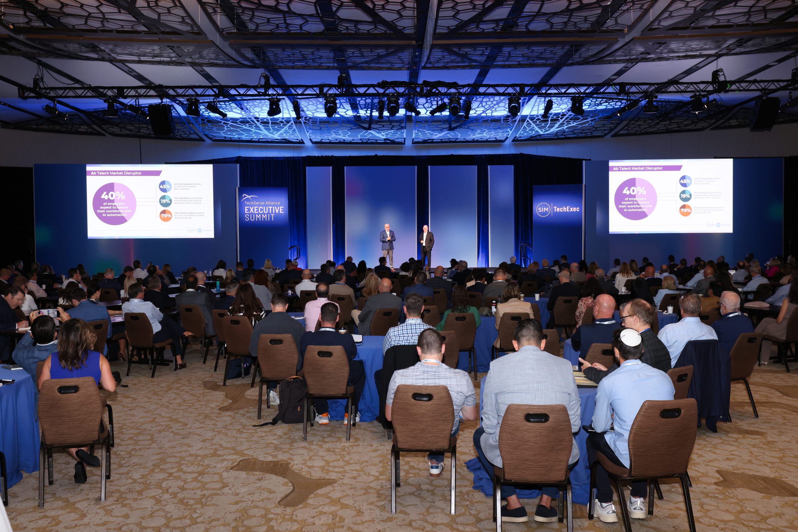 Executive Summit 8 A large audience sits in a conference room facing a stage where two speakers present. Two projection screens display charts and text. The event is labeled “Executive Summit.” The room is brightly lit with a modern ceiling design.