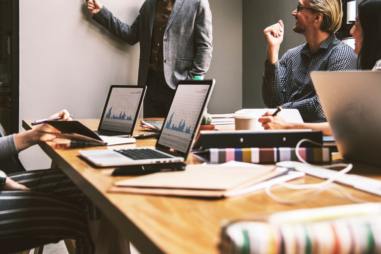Four people are in a meeting room, with one standing and pointing at a whiteboard featuring market data. Three others sit around a table with laptops displaying graphs, notebooks, and folders. The setting appears collaborative and professional.