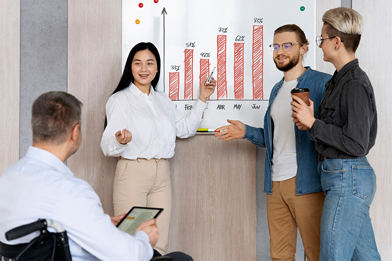 Three people stand by a whiteboard with a red bar graph, discussing data and ways to optimize IT staffing in a competitive marketplace, while a fourth person in a wheelchair listens and holds a tablet. The atmosphere appears collaborative and engaged.