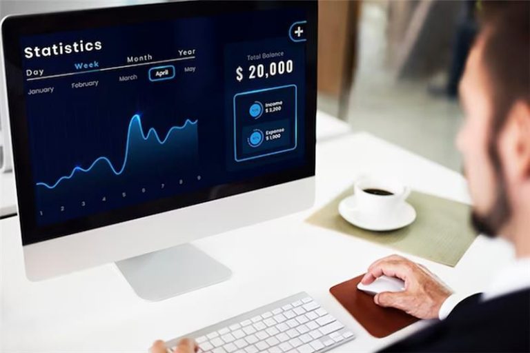 A man in business attire uses a computer displaying a statistics dashboard with a line graph, monthly data, and a total balance of $20,000—showcasing how AI in Sales can drive real results. A cup of coffee and keyboard are on the desk.