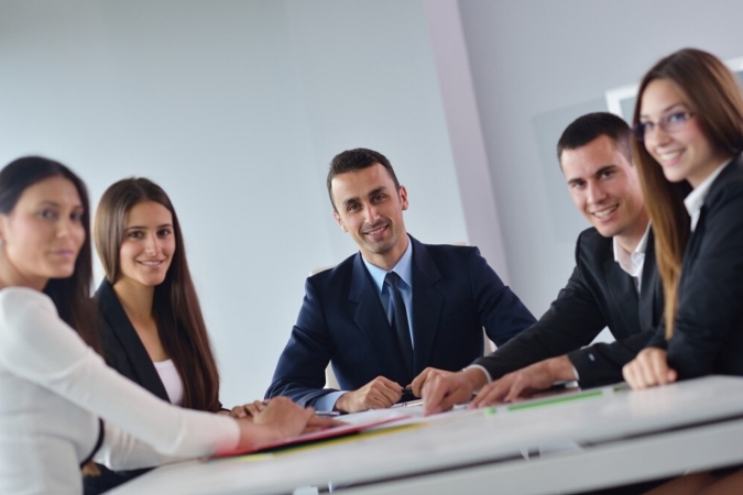 Five professionally dressed people sit around a conference table, smiling and looking toward the camera, suggesting an Executive Roundtable or teamwork setting in a modern office environment.