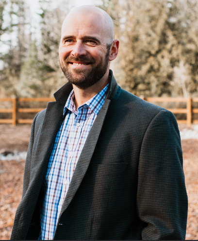 A smiling bald man with a beard, wearing a checked shirt and dark blazer, stands outdoors in front of a wooden fence and trees—an approachable face for Tech Consulting and the Expert Network at TechServe.