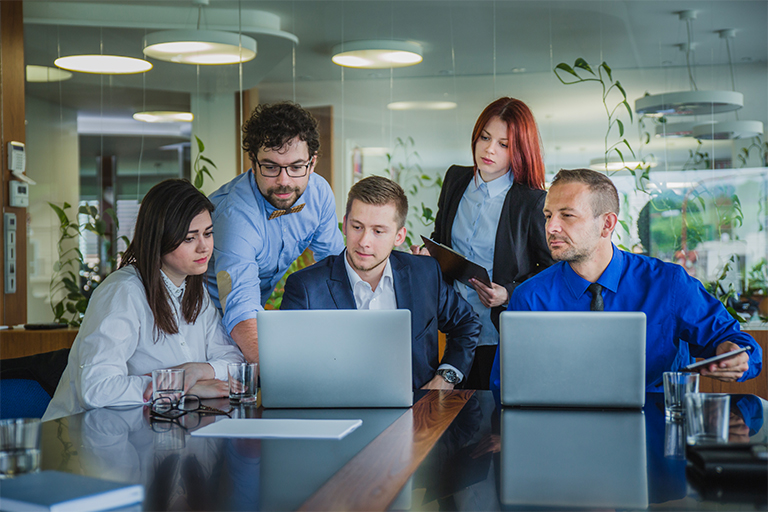Five people in business attire sit and stand around a table, focused on two open laptops, discussing IT staffing needs in a modern office with glass walls and plants in the background.