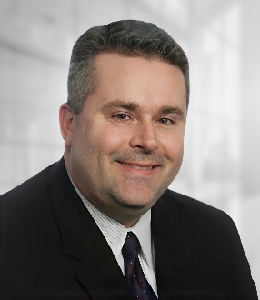 A man with short gray hair wearing a dark suit, white shirt, and patterned tie, smiles at the camera against a blurred office background, reflecting his professionalism as part of the TechServe Expert Network.