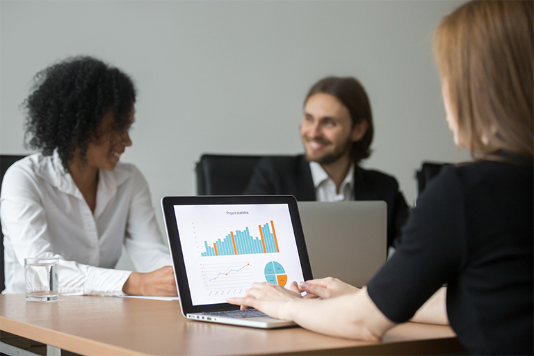 Three people sit at a conference table with laptops. In the foreground, a woman displays tech investments charts on her screen, while two colleagues smile and discuss recruiting results in the background.