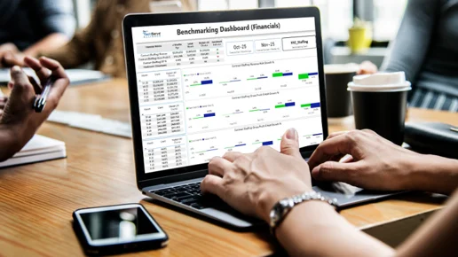 A person uses a laptop displaying a financial benchmarking dashboard in an office setting, with others nearby, papers, a smartphone, and a coffee cup on the wooden table.
