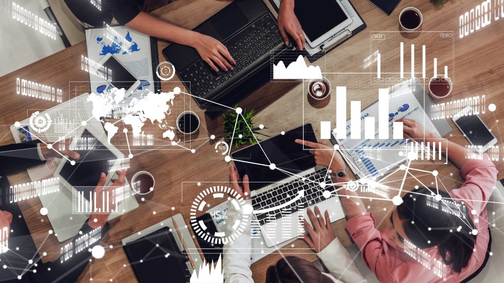 Breaking Down Silos: Why Sales, Marketing, and Technology Must Act as One Revenue Engine 1 Overhead view of people working at a table with laptops, tablets, and papers, surrounded by digital graphs, charts, and data visualizations representing analytics, sales performance, and business teamwork.