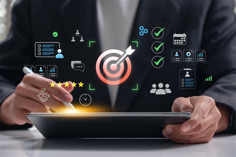 A person in a suit uses a tablet with a stylus, surrounded by digital icons representing data, analytics, teamwork, communication, checklist tasks, and a large central target with an arrow.