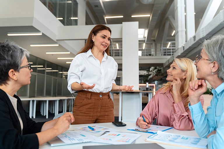 A woman founder stands and speaks to three colleagues seated at a table covered with papers and charts in a modern office setting, demonstrating leadership as the group engages in discussion or a meeting.
