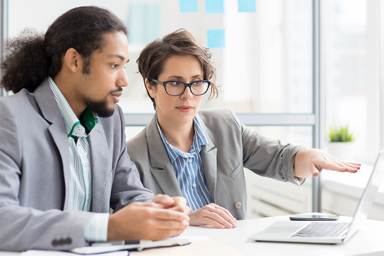 Two professionals in business attire sit at a desk, focused on a laptop screen discussing IT staffing. One gestures toward the screen while the other listens attentively in a bright office setting with large windows in the background.