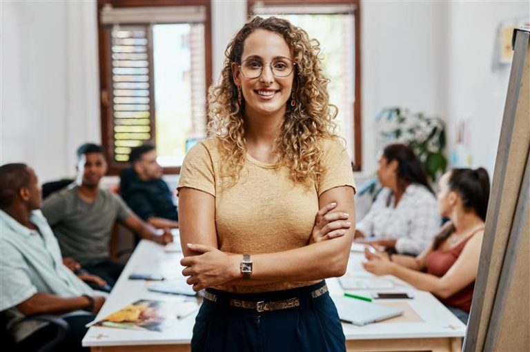 A woman with curly hair and glasses stands confidently with arms crossed, embodying leadership, in front of a conference table where four people are having a meeting in a bright, modern office.