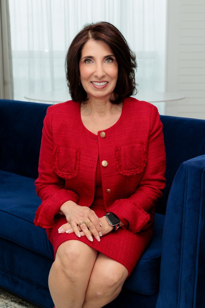 A woman with shoulder-length dark hair, wearing a red textured blazer and skirt, sits on a blue velvet couch. Smiling with her hands folded on her knee and a smartwatch on her wrist, she embodies the professionalism of the TechServe expert network.