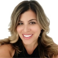 Jessica Sellitto, a woman with long, wavy, light brown hair, smiles at the camera. She is wearing a black sleeveless top and small earrings, with a white background behind her.