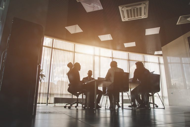 Silhouetted group of Staffing Leaders sitting around a conference table in a modern office with large windows, sunlight streaming in, and ceiling lights visible above.