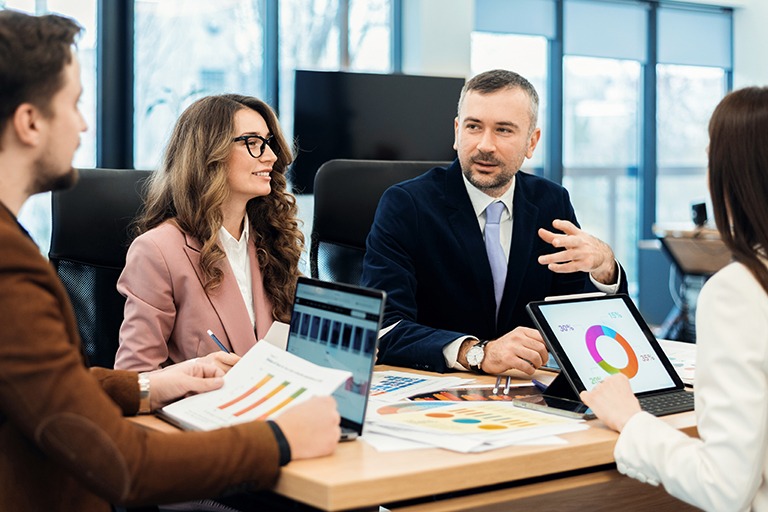 Four people in business attire sit around a conference table discussing charts and graphs on paper and digital devices, suggesting a professional staffing sales leadership meeting or presentation in a modern office setting.