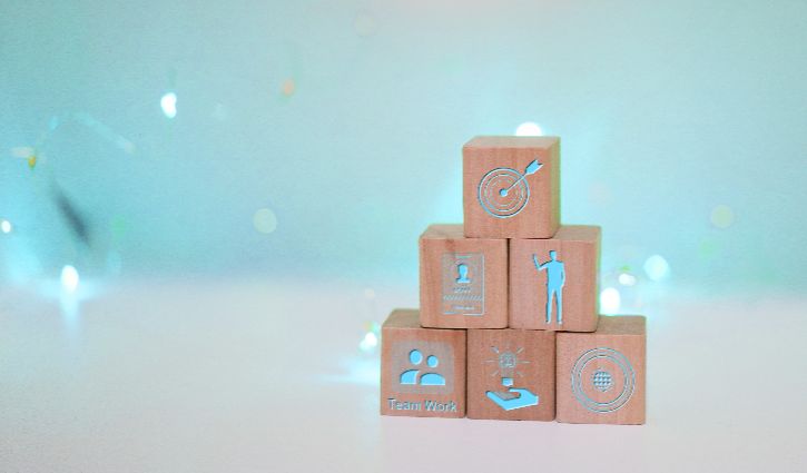 A pyramid of five wooden blocks with icons for teamwork, personal profile, a hand holding a gear, a target, and a person pointing at a board—symbolizing sales leadership and daily habits—against a light blue background with soft lights.